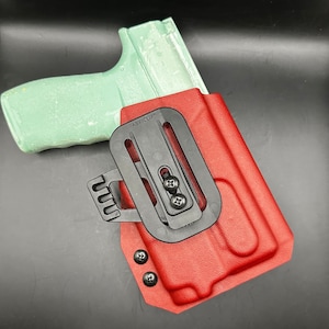 May include: A red leather holster with a black fabric clip and a black plastic clip. The holster has two black screws on the side and two black screws on the bottom. The text "FABRICLIP" is printed on the black fabric clip.