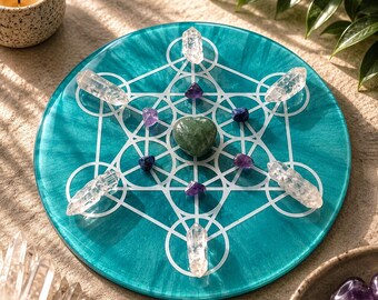 May include: A circular teal resin plate featuring a white Metatron's Cube geometric pattern. The plate is adorned with six clear quartz points, several small purple amethyst tumbled stones, and a central green heart-shaped stone.