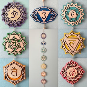 May include: A collection of seven laser-cut wooden wall hangings representing chakra symbols, each featuring distinct geometric shapes and vibrant colours including purple, blue, teal, green, yellow, orange, and red, displayed individually and as a vertical string.