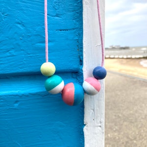 Può includere: Una collana colorata con perline di legno dipinte nei colori giallo, turchese, rosa, bianco e blu. Le perline sono infilate su un cordino rosa. La collana è esposta su uno sfondo blu e bianco.