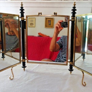 May include: A three-sided brass and black vanity mirror with a decorative design on the top of each side. The mirror is standing on a table with a white tablecloth.