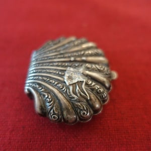 Puede incluir: Una caja de alhajas de plata con forma de concha, con intrincados diseños florales. La caja está cerrada y tiene un diseño detallado en el exterior.