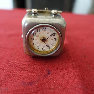 Puede incluir: Pequeño reloj de viaje plateado vintage, de forma cuadrada, con esfera redonda. La esfera tiene números y agujas negros sobre fondo crema. Colocado sobre una superficie roja.