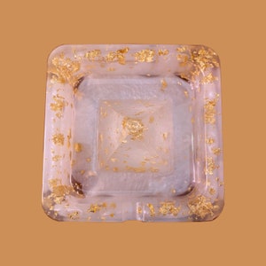 May include: A square, translucent resin ashtray featuring a pearlescent white base embedded with scattered gold leaf flakes. A small, matching pyramid-shaped resin object with gold leaf accents sits centered within the square basin of the ashtray.