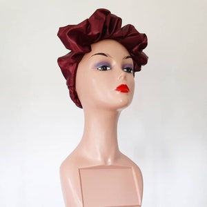 May include: A burgundy satin hair bonnet with a gathered design. The bonnet is worn on a mannequin head with a neutral makeup look and red lipstick.