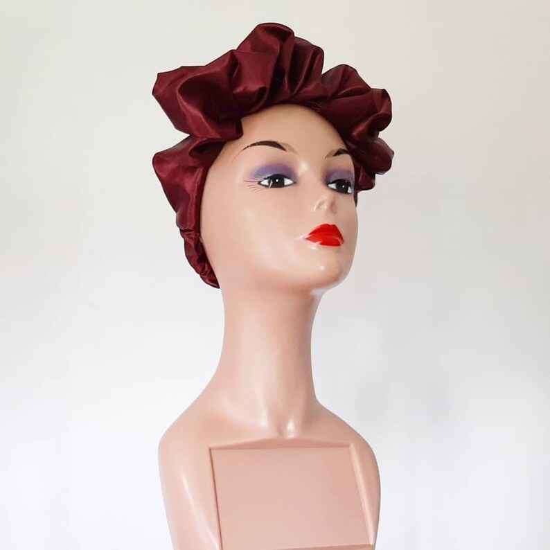 May include: A burgundy satin hair bonnet with a gathered design. The bonnet is worn on a mannequin head with a neutral makeup look and red lipstick.