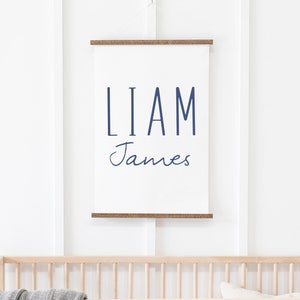 May include: A white canvas print with the names "Liam James" in navy blue lettering. The print is hanging on a white wall above a wooden crib.