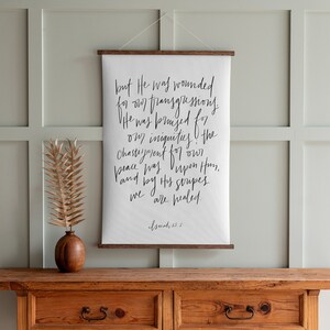 May include: A white canvas wall hanging with a black handwritten quote from Isaiah 53:5. The quote reads "but He was wounded for our transgressions. He was bruised for our iniquities. The chastisement for our peace was upon Him, and by His stripes we are healed." The quote is framed with a dark wood frame and is hanging on a light green wall.