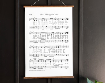 May include: A black and white printable sheet music poster with the title "The Old Rugged Cross" and the song lyrics printed on it. The poster is framed with a wooden frame and hanging on a black wall.