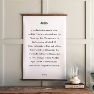 May include: A white canvas wall hanging with a wooden frame and the text "JOHN" in black. The text is a bible verse from John 1:1-5, "In the beginning was the Word, and the Word was with God, and the Word was God. The same was in the beginning with God. All things were made by him; and without him was not any thing made that was made. In him was life; and the life was the light of men. And the light shineth in darkness; and the darkness comprehended it not. 1:1-5"