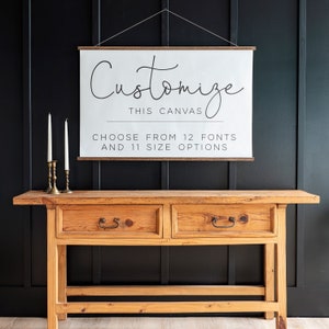 May include: A white canvas with a wooden frame hanging on a black wall. The canvas has the text "Customize this canvas. Choose from 12 fonts and 11 size options." printed on it. A wooden table with two drawers is in the foreground.