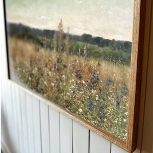 May include: A framed painting of a field of wildflowers with a blurred background of trees and a cloudy sky. The wildflowers are mostly white and yellow with some blue flowers.