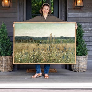 May include: A rectangular landscape wall art print featuring a field of wildflowers in shades of green, yellow, and blue under a pale sky. The artwork includes cursive white text that reads, Consider the lilies of the field, how they grow.