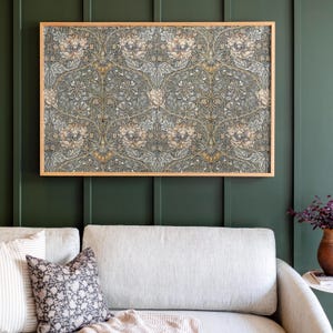 Op de afbeelding: Een ingelijste print van een vintage bloemenpatroon in tinten groen, bruin en wit. De print hangt aan een donkergroene muur boven een lichtgrijze bank.