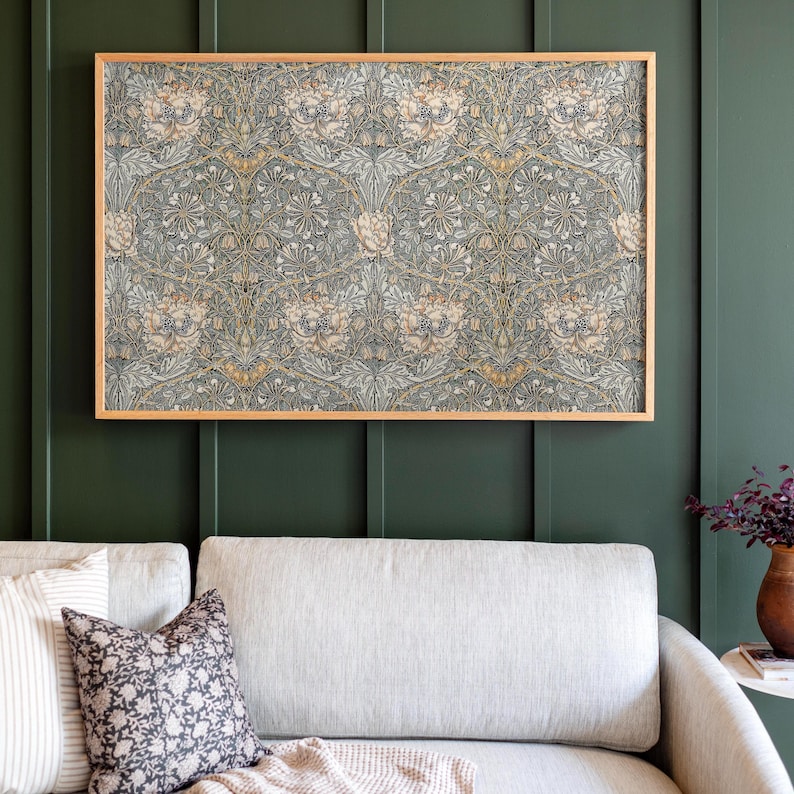 Op de afbeelding: Een ingelijste print van een vintage bloemenpatroon in tinten groen, bruin en wit. De print hangt aan een donkergroene muur boven een lichtgrijze bank.