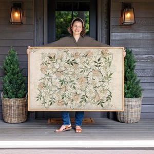 May include: A large rectangular framed wall art piece featuring a repeating botanical pattern of light beige flowers and dark green leaves on a cream-colored background. The artwork is enclosed in a simple, light-toned wooden frame.