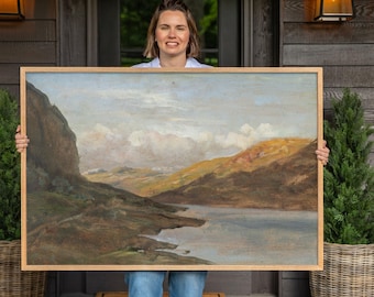 May include: A rectangular landscape painting in a light wood frame depicting a serene lake surrounded by rolling hills and mountains under a cloudy sky. The artwork features earthy tones of brown, green, and muted blue, capturing a natural vista.