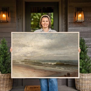 May include: A rectangular framed landscape painting depicting a coastal scene with a sandy beach, gentle ocean waves, and a cloudy sky. The artwork features muted tones of beige, grey, and blue, showcasing a calm shoreline with small rocks.