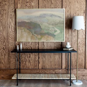 May include: A black console table with a woven shelf and a gold-framed abstract painting above it. The table is against a wood-panelled wall with a floor lamp to the right.
