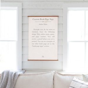 May include: A white sign with black text that reads "Custom Book Page Sign" and "Author Name". The text continues with instructions on how to customize a book page sign. The sign is hanging on a white wall with a wooden texture. The text at the bottom of the sign reads "55."