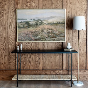 May include: A black console table with a woven shelf and two small white ceramic vases on top. The table is in front of a wood-paneled wall with a framed landscape painting above it. A white lamp with a gold base is on the right side of the table.