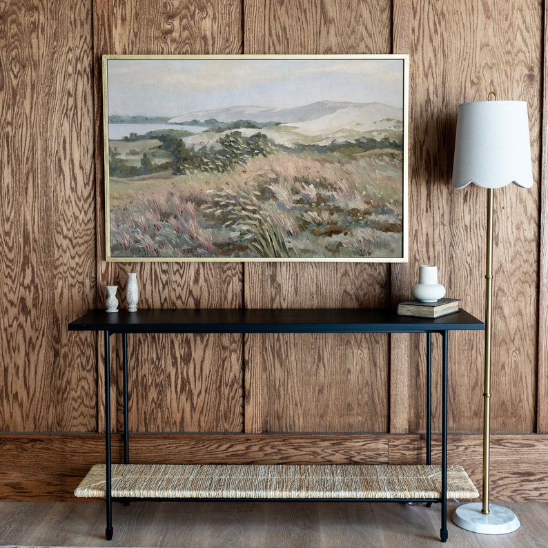 May include: A black console table with a woven shelf and two small white ceramic vases on top. The table is in front of a wood-paneled wall with a framed landscape painting above it. A white lamp with a gold base is on the right side of the table.