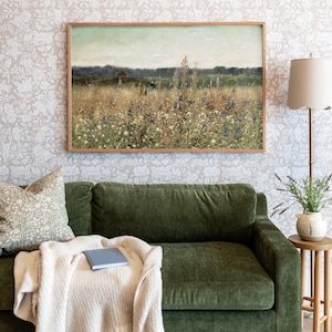 May include: A green velvet sofa with a throw blanket and a book on it. A framed landscape painting of a field of wildflowers hangs above the sofa. A lamp with a white shade sits on a table next to the sofa.