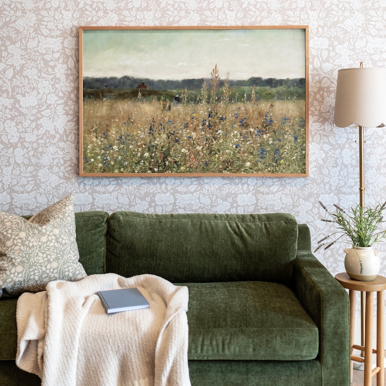 May include: A green velvet sofa with a throw blanket and a book on it. A framed landscape painting of a field of wildflowers hangs above the sofa. A lamp with a white shade sits on a table next to the sofa.