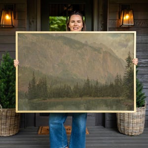 May include: A rectangular landscape painting in a thin gold frame depicting a calm lake at the base of a large, steep mountain range. The scene features a dense forest of dark green pine trees along the shoreline under a muted, cloudy sky.