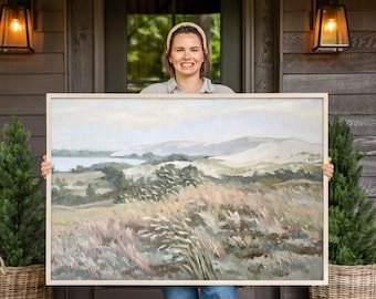 May include: A rectangular landscape painting in a light wood frame depicting a grassy field in the foreground with rolling hills and a body of water in the background. The artwork features a muted color palette of soft greens, tans, and pale blues.