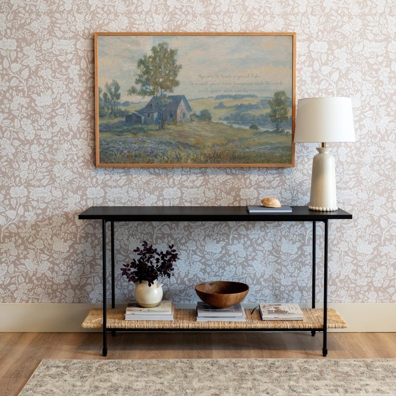 May include: A black console table with a woven shelf, holding a white vase with dark purple stems, a wooden bowl, and books. A framed landscape painting hangs above the table, and a white lamp with a beaded base sits on the right.