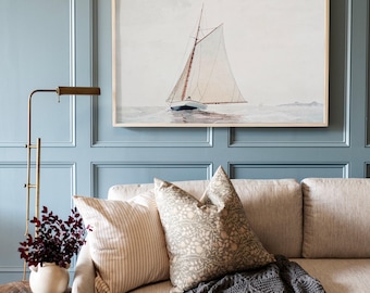 May include: A framed horizontal wall art print featuring a sailboat with white sails on calm water. The artwork is displayed above a beige sofa with a striped cushion, a floral patterned cushion, and a dark grey knit throw blanket.