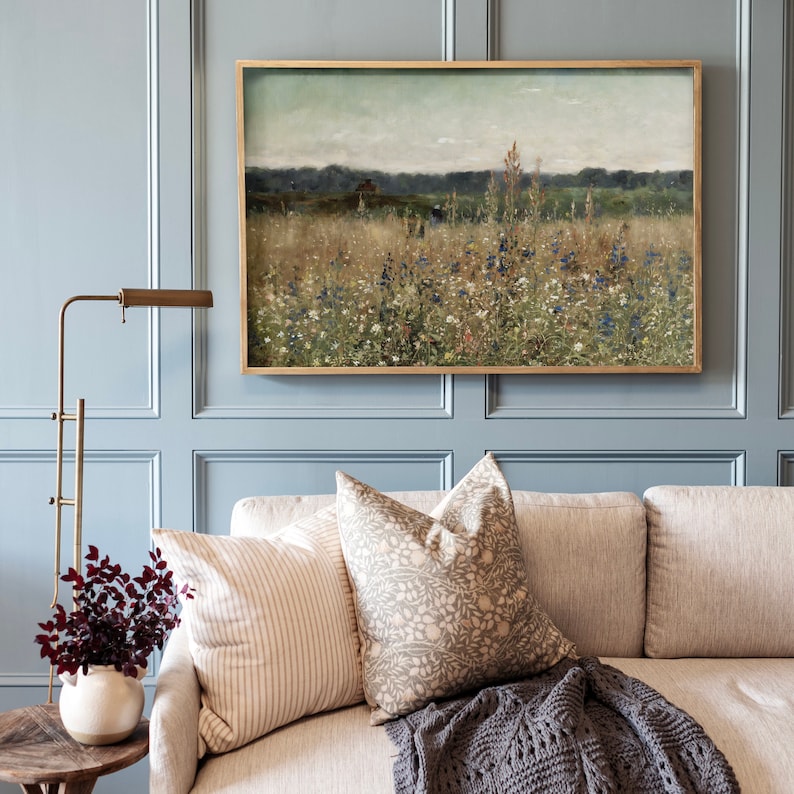 May include: A light blue sofa with a gray knitted throw blanket and two throw pillows. One pillow is a beige and brown floral pattern and the other is a white and beige striped pattern. A framed painting of a field of wildflowers hangs above the sofa.