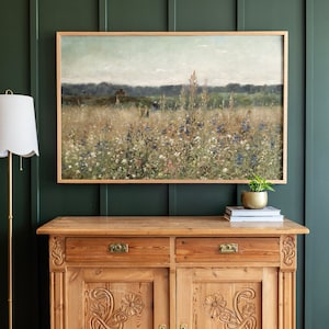 May include: A framed landscape painting depicting a field of wildflowers in shades of green, blue, and white. The painting is hanging on a dark green wall above a wooden cabinet with carved details.