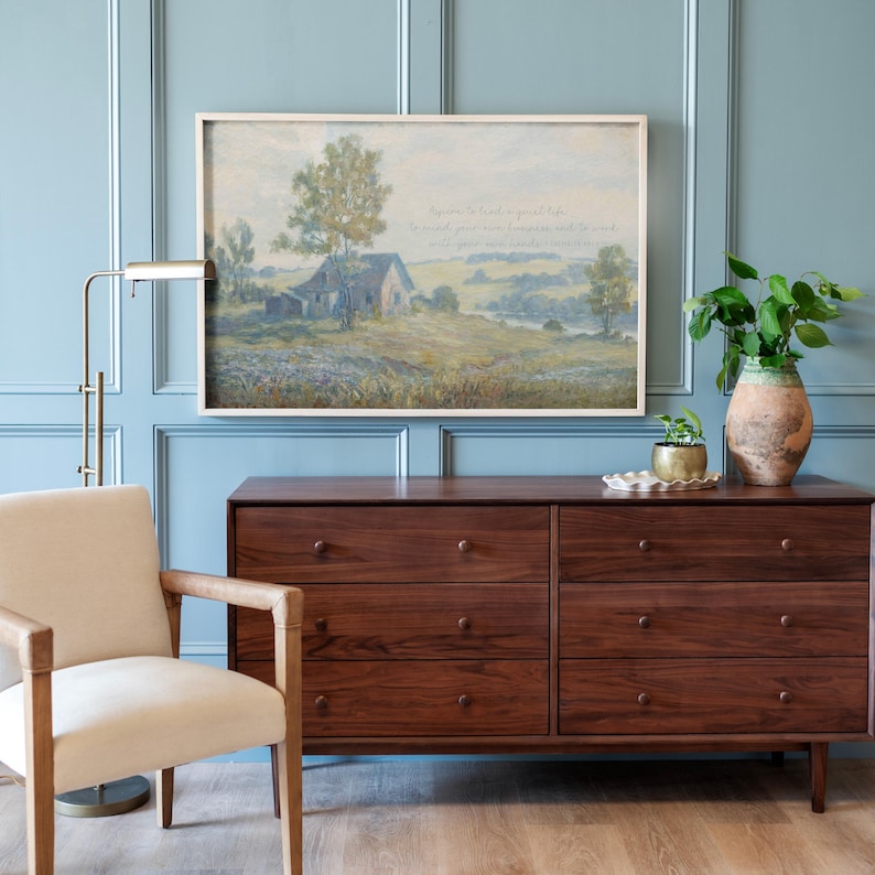 May include: A framed landscape painting with a light-colored frame hangs above a dark wood dresser. The painting depicts a rural scene with a house, trees, and fields. A beige chair and a brass lamp are to the left of the dresser. A vase with greenery sits on the dresser.