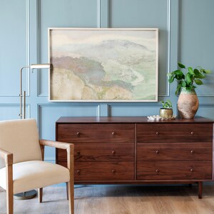 May include: A light blue wall with a framed abstract landscape painting above a dark brown dresser with six drawers. A white armchair with wooden arms is to the left of the dresser. A gold floor lamp is behind the armchair. A potted plant is on the right side of the dresser.