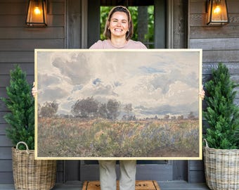 May include: A large rectangular landscape painting in a thin gold frame featuring a field of wildflowers under a cloudy sky. The artwork depicts a natural scene with muted tones of green, brown, and blue, showcasing a vast horizon with trees.