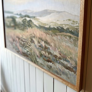 May include: A framed painting of a landscape with a view of rolling hills and a field of tall grass. The painting is done in soft, muted colors, with shades of green, brown, and blue. The frame is a simple, natural wood.