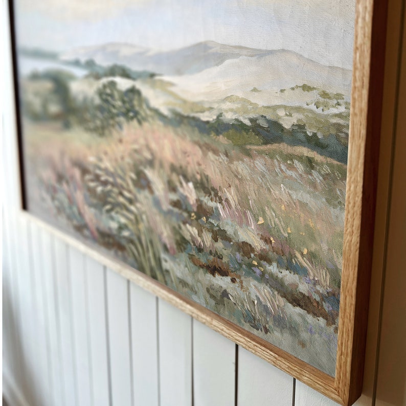 May include: A framed painting of a landscape with a view of rolling hills and a field of tall grass. The painting is done in soft, muted colors, with shades of green, brown, and blue. The frame is a simple, natural wood.