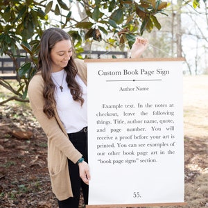 May include: A white sign with a wooden frame hangs from a string. The sign says "Custom Book Page Sign" and includes instructions for ordering a custom book page sign. The text says "Example text. In the notes at checkout, leave the following things. Title, author name, quote, and page number. You will receive a proof before your art is printed. You can see examples of our other book page art in the "book page signs" section."
