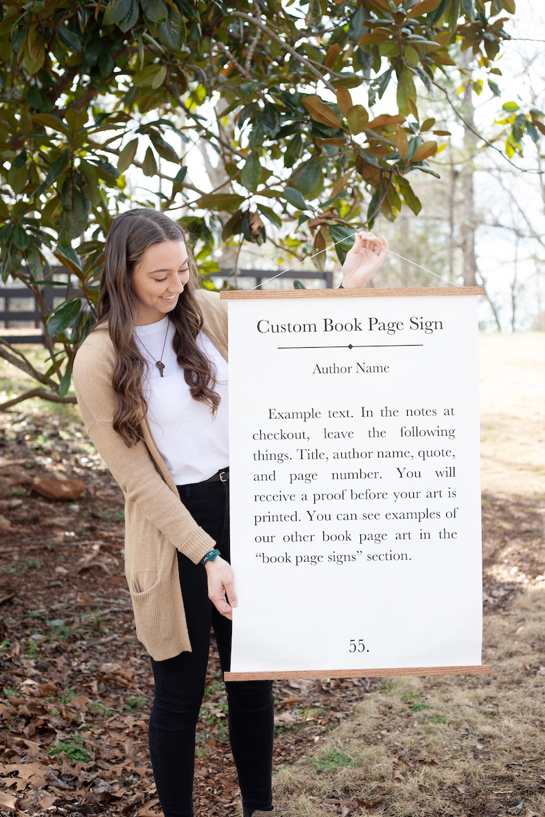 May include: A white sign with a wooden frame hangs from a string. The sign says "Custom Book Page Sign" and includes instructions for ordering a custom book page sign. The text says "Example text. In the notes at checkout, leave the following things. Title, author name, quote, and page number. You will receive a proof before your art is printed. You can see examples of our other book page art in the "book page signs" section."