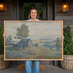 May include: A framed landscape painting with a rustic farmhouse and a quote. The artwork features a muted color palette of blues, greens, and browns. The wooden frame adds a natural touch to the artwork. The painting is held by a person.