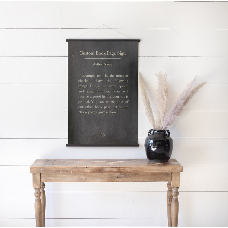 May include: Black and white custom book page sign with the text "Custom Book Page Sign" and "Author Name". The sign is hanging on a white wall above a wooden table with a black vase and dried flowers.