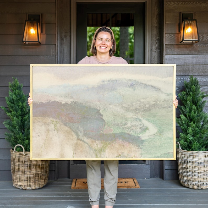 May include: A woman is holding a large framed abstract painting with a gold frame. The painting depicts a landscape with shades of green, blue, and brown.