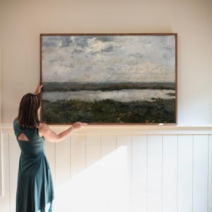 May include: A rectangular landscape painting in a dark wood frame featuring a cloudy sky over a dark green marsh and water. The artwork includes cursive text written across the centre of the sky area, depicting a natural outdoor scene.