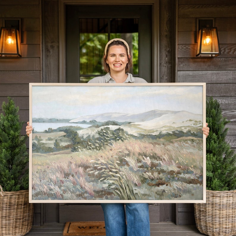 May include: A woman holds a framed landscape painting depicting a grassy field with rolling hills in the background. The painting has a light blue, green, and brown color scheme.