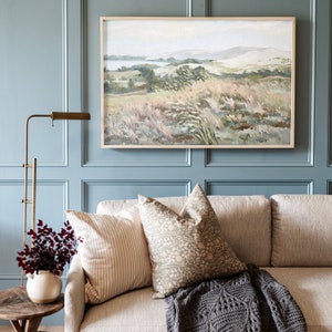 May include: A living room interior with a light blue wall, a couch with throw pillows and a blanket, a floor lamp, and a framed painting of a landscape with a body of water in the distance.
