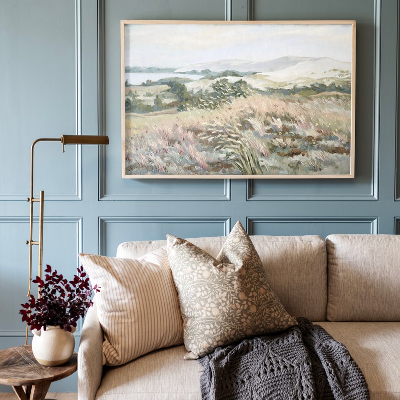 May include: A living room interior with a light blue wall, a couch with throw pillows and a blanket, a floor lamp, and a framed painting of a landscape with a body of water in the distance.