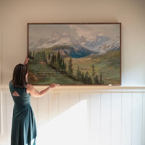 May include: A framed landscape painting depicting a mountain range with snow-capped peaks, a valley, and evergreen trees. The artwork has a wooden frame and includes text in the lower left corner. The painting is displayed on a white wall.