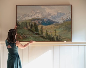 May include: A framed landscape painting depicting a mountain range with snow-capped peaks, a valley, and evergreen trees. The artwork has a wooden frame and includes text in the lower left corner. The painting is displayed on a white wall.